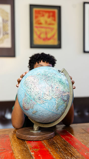 a young boy stands at a wood table with his face obscured by a globe. He has one hand on either side of the globe and his dark, curly hair peeks out over the top of the globe.