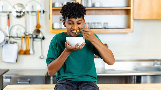 A young boy with dark curly hair wears a green t-shirt and sits on a wooden counter in a hostel kitchen. He holds a large face while spooning colorful cereal into his mouth.