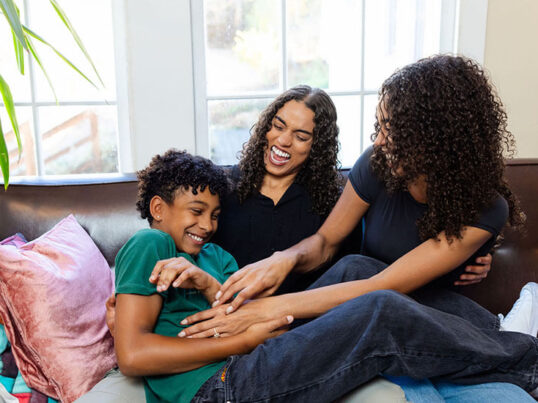 A family relaxes in a hostel lounge. Two moms with curly hair and black t-shirts smile while tickling their young son. The son wears a green t-shirt and laughs while leaning back on a comfortable sofa in a hostel lounge.