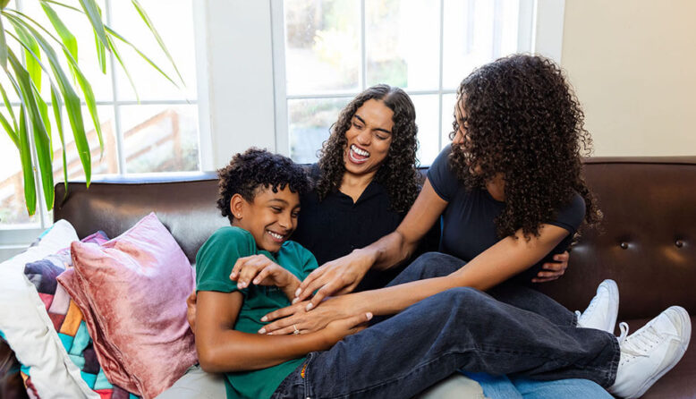 A family relaxes in a hostel lounge. Two moms with curly hair and black t-shirts smile while tickling their young son. The son wears a green t-shirt and laughs while leaning back on a comfortable sofa in a hostel lounge.
