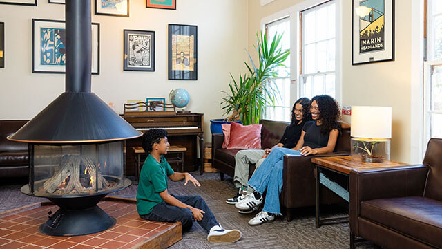 in a cozy hostel lounge with a central fire place, two moms and their young son sit talking.