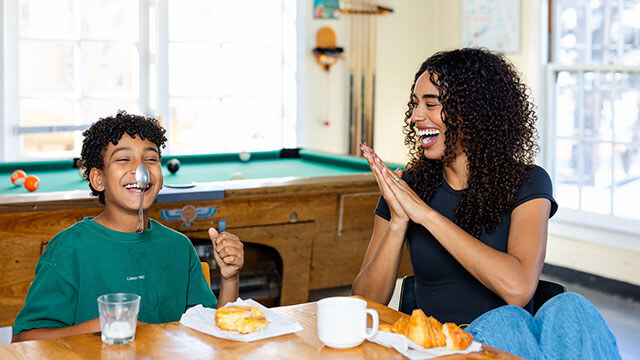 A young boy in a green t-shirt balances a spoon on his nose while sitting at the breakfast table in a hostel. There is a pool table in the background. The boy's mother, who wears a black t-shirt and jeans and has dark curly hair, smiles and claps for her son.