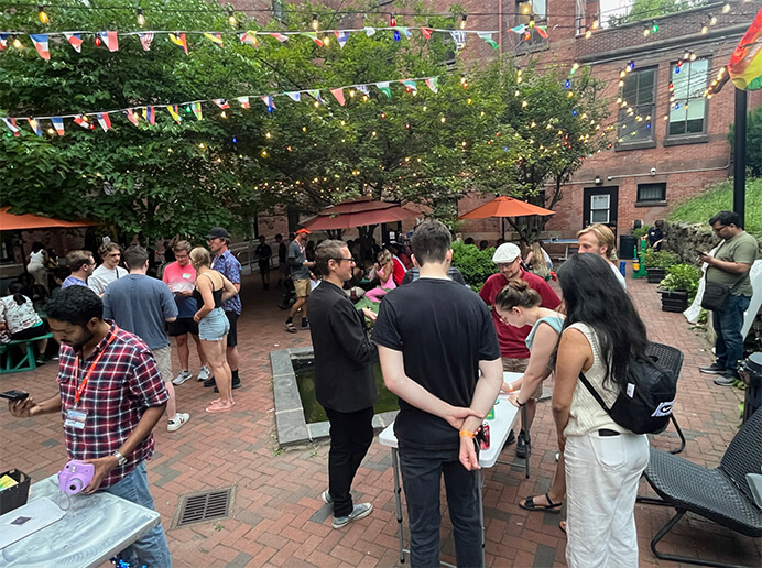 Travelers outside at dusk in the back garden at HI New York City hostel. There are small international flags draped overhead on the brick patio, with picnic tables, large red umbrellas, and trees.