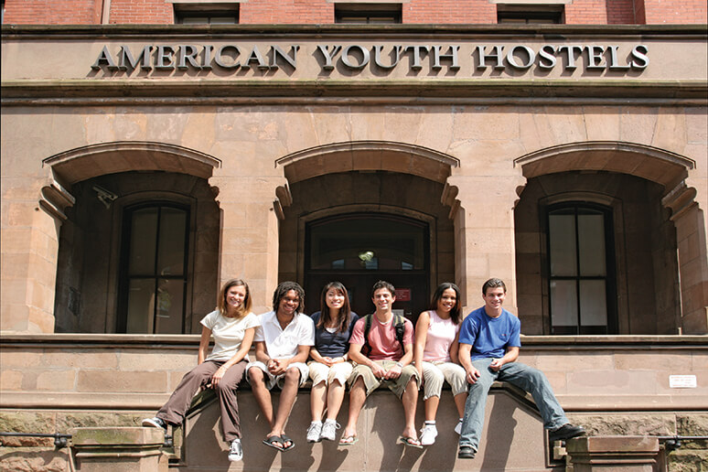 Six young travelers sit on a wall in front of thee stone arches making up the entryway to a huge brick building.