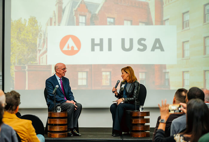 Two people sit facing each other on a stage. Behind them, a large screen shows the HI USA logo with a photo of HI New York City Hostel behind it. In the foreground, a crowd of people sit watching.