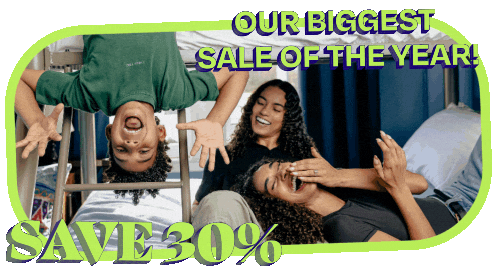 Two moms on the lower level of a bunk bed laugh as their young son hangs upside down from the top bunk making a silly face. The moms are both wearing black shirts, and the son is in a green t-shirt. A text overlay reads 