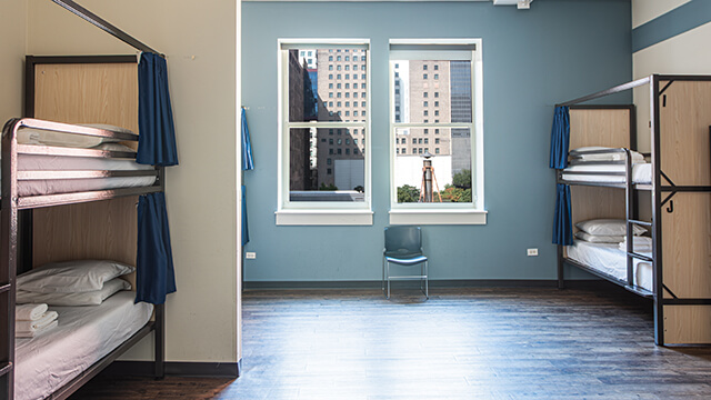 A large dorm room at a HI USA hostel. There are wood floors, pale blue walls, and several windows letting in natural light. Each bunk bed has its own privacy curtain.