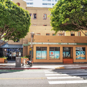 The exterior of HI Los Angeles Santa Monica hostel on a sunny day. The photo is taken from across the street, with two bush trees framing the exterior of the building.