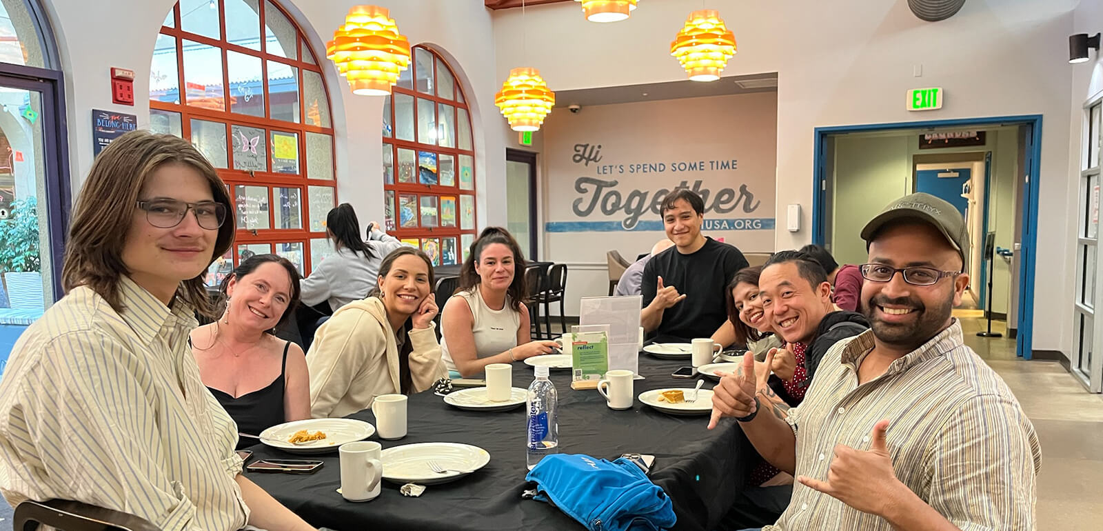A group of guests at HI Los Angeles Santa Monica hostel sit around a dining table in the hostel dining room enjoying a group meal during a hostel-organized event.