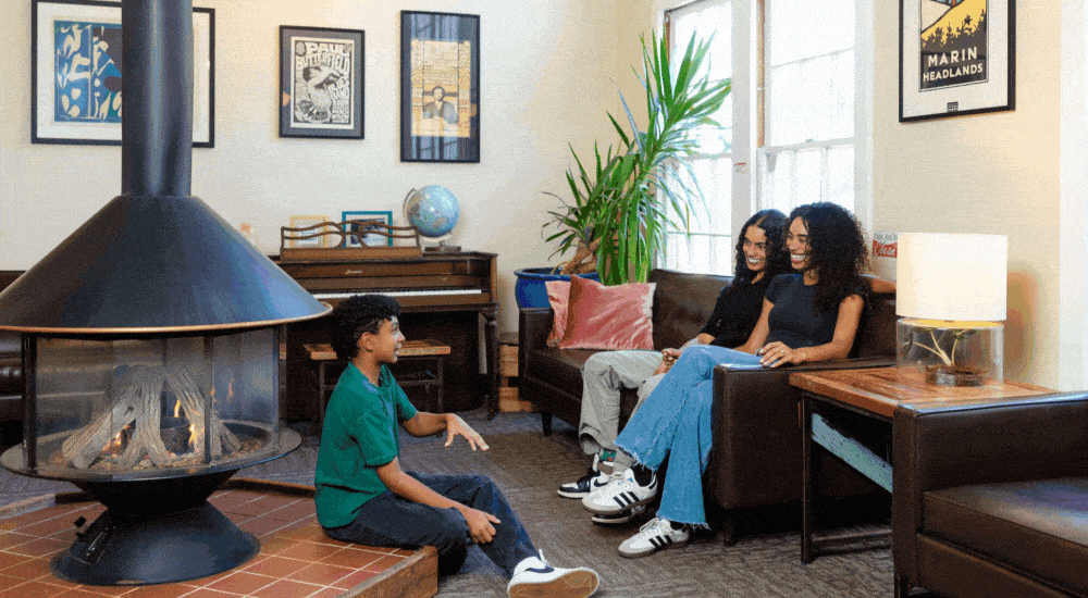 Two moms and their young son sit by a fireplace in a cozy hostel lounge.