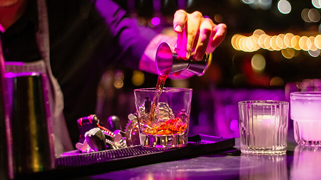 A hand pouring a shot of alcohol into a glass with ice under pink lighting.