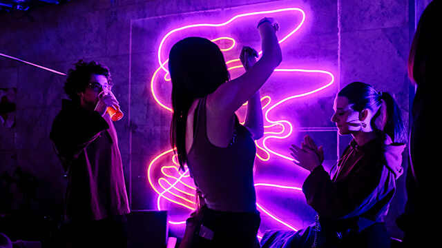 a silhouette of a young traveler is seen dancing against neon pink and purple lights at a party hostel.