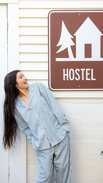 A young traveler with long black hair wearing green-and-white-striped pajamas stands under a brown-and-wite sign reading "hostel" with a graphic of a hut and tree on it. She leans to the side and looks up at the sign.