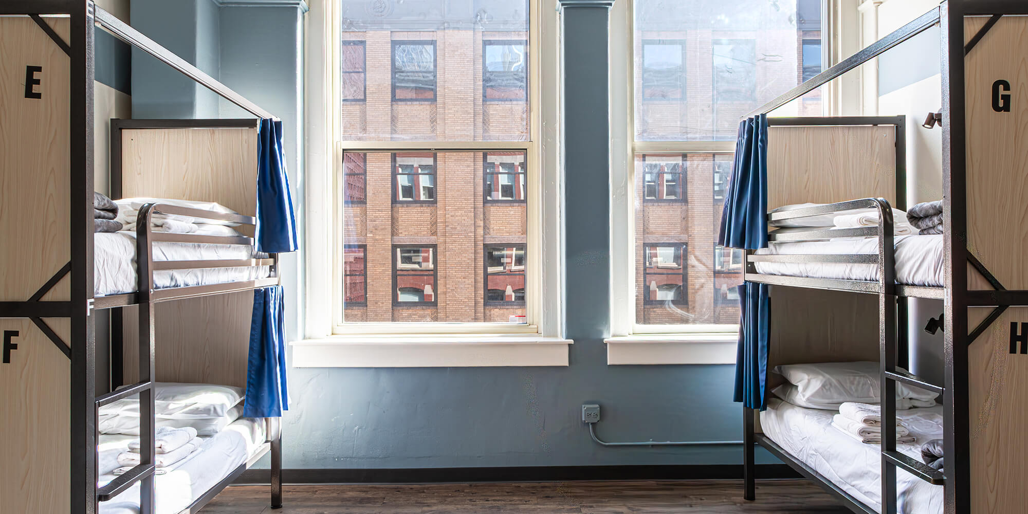 A modern dorm room at HI Chicago hostel featuring sturdy bunk beds with privacy curtains, pale blue walls, and windows overlooking the city letting in lots of natural light.