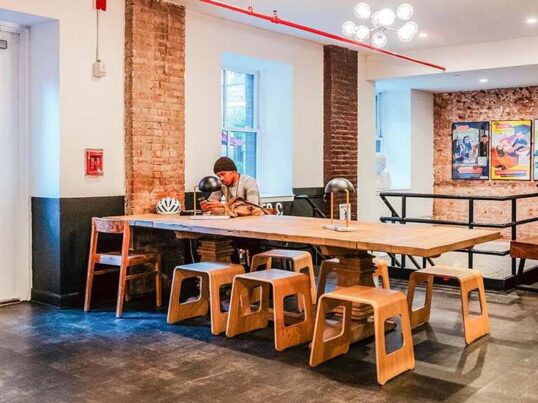 Digital nomads working at a long wooden coworking table in a HI New York City hostel common area, featuring exposed brick walls and natural light.