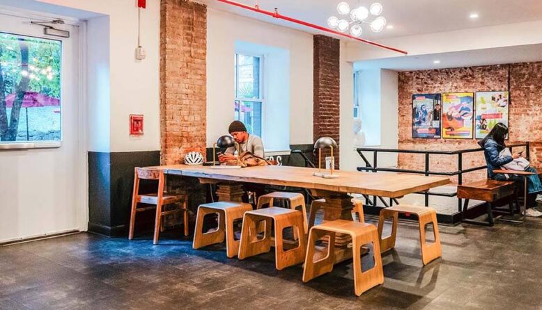 Digital nomads working at a long wooden coworking table in a HI New York City hostel common area, featuring exposed brick walls and natural light.