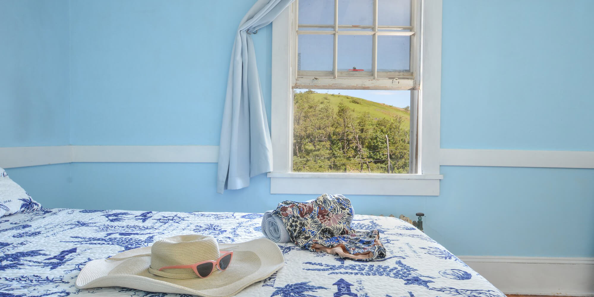 An airy and light-filled private room at HI Truro hostel. There is a queen-sized bed with fresh blue-and-white linens. On top of the bed is a wide-brimmed sun hat, pink-framed sunglasses, and a floral-printed swimsuit. The open window has a view outside.