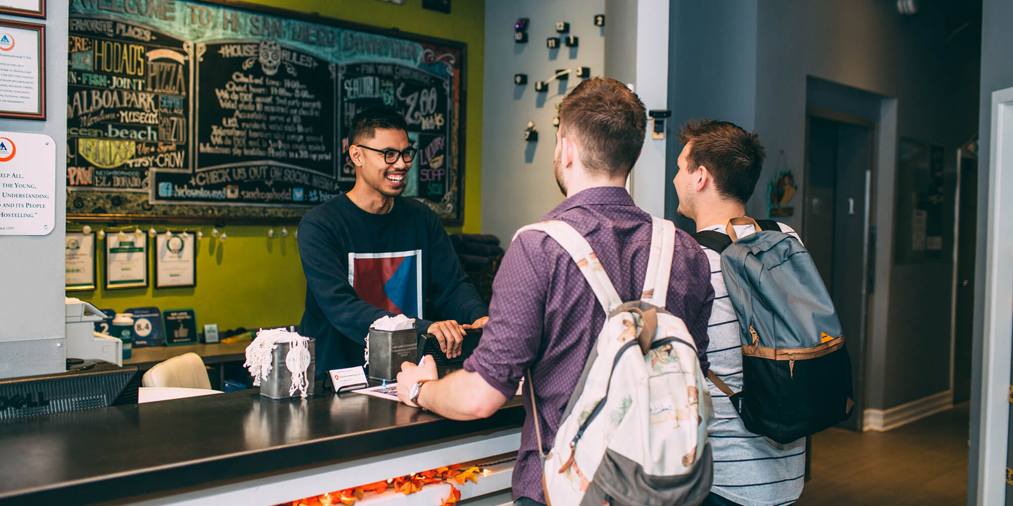 A friendly front desk agent at HI San Diego Downtown hostel greets two guests wearing backpacks