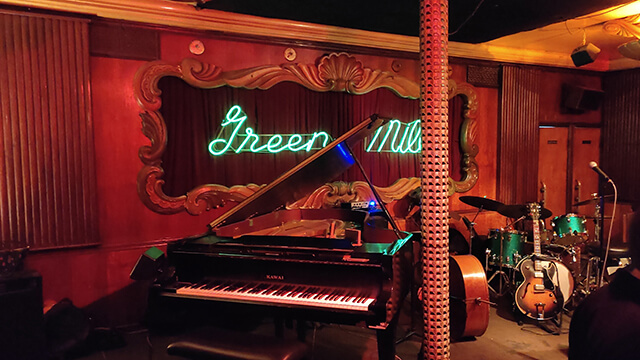 The interior jazz stage at The Green Mill in Chicago, featuring a black grand piano, a drum set, and a double bass set against a warm wood-paneled wall with a glowing green neon "Green Mill" sign inside an ornate gold frame.
