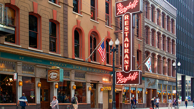A street-level view of The Berghoff Restaurant in downtown Chicago, featuring its iconic red vertical neon sign and historic brownstone facade with arched windows on Adams Street near the Route 66 starting point.