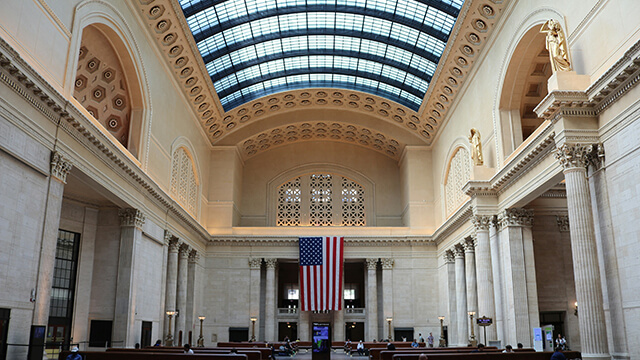 The massive interior of the Great Hall at Chicago Union Station, featuring towering Corinthian columns, a huge vaulted skylight, and a large American flag hanging from the far wall, representing the historic 1925 landmark near the start of Route 66.