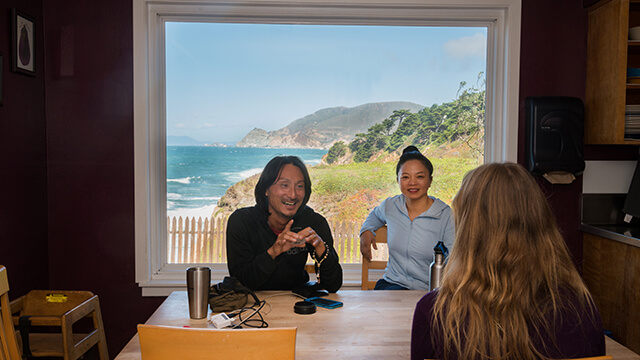 Guests sitting at a communal table in the Sea Lodge at HI Point Montara Lighthouse, featuring a large picture window with stunning Pacific Ocean views.