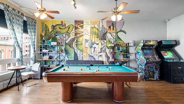 Vibrant game room at HI San Diego Downtown featuring a pool table, vintage arcade games, and a colorful local art mural.