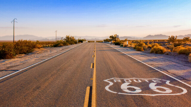 A long, straight stretch of historic Route 66 stretching into a desert horizon at sunset, with a large white Route 66 logo painted directly on the asphalt in the foreground.