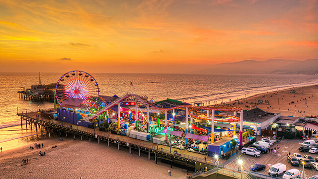 High-angle sunset view of the Santa Monica Pier and Pacific Park amusement park, featuring a glowing Ferris wheel and roller coaster over the ocean with a vibrant orange and pink sky.