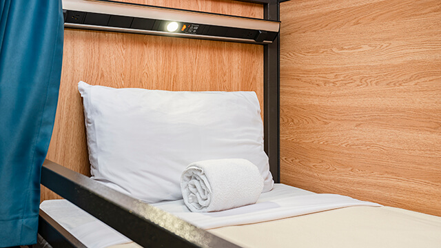 Detail of a comfortable bunk bed at a HI USA hostel with a pillow and towel, reading light, charging ports, and a blue privacy curtain.